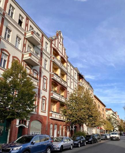 Straßenszene in Berlin-Wedding mit mehrstöckigem Altbau in rotem Backstein und hell verputzten Fassaden, Balkonen sowie geparkten Autos und Bäumen am Straßenrand.

