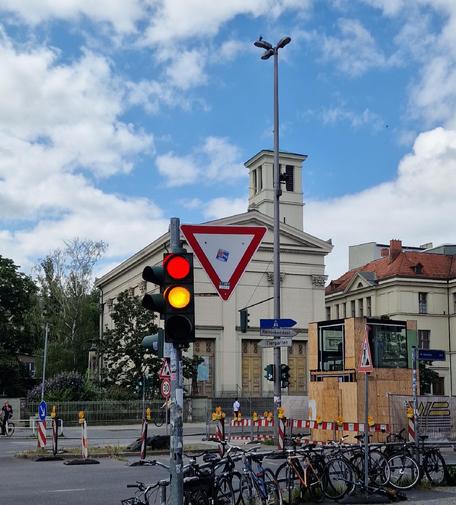 Ampel mit Rot- und Gelblicht an einer Straßenkreuzung in Berlin-Wedding, im Hintergrund die Nazarethkirche und Baustellenabsperrungen, davor mehrere abgestellte Fahrräder.

