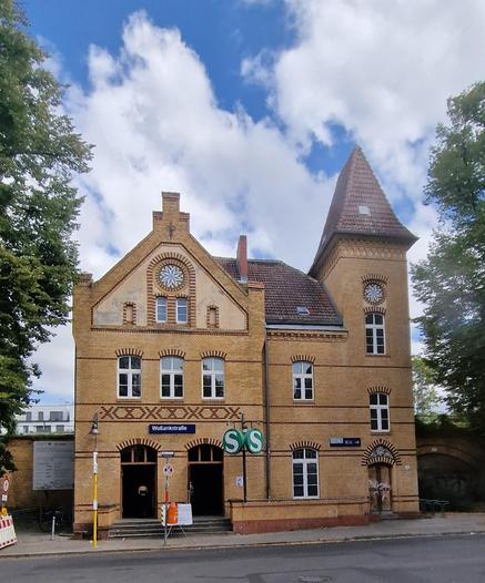 Historisches Backsteingebäude des S-Bahnhofs Wollankstraße in Berlin-Wedding, mit spitzem Turm, Rundbogenfenstern und den grünen S-Bahn-Schildern am Eingang.

