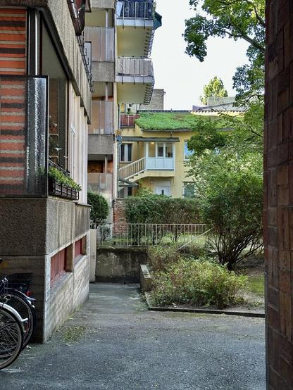 Hinterhofszene mit Mehrfamilienhäusern, Balkonen und einem begrünten Dach; im Vordergrund Fahrräder und ein kleiner Gartenbereich mit Sträuchern.

