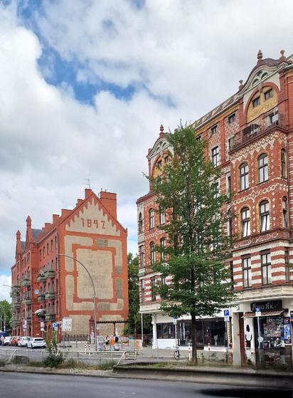 Auf dem Foto sind zwei historische Mietshäuser aus Backstein zu sehen, die sich durch reich verzierte Fassaden auszeichnen. Am linken Gebäude ist deutlich die Jahreszahl 1892 als Baujahr angebracht. Die Architektur mit Ziergiebeln, Balkonen und dekorativen Mustern im Mauerwerk verweist auf die Gründerzeit. Vor den Gebäuden verlaufen eine Straße und Gehwege, im Erdgeschoss des rechten Hauses befinden sich Geschäfte, darunter ein Fotoladen. Ein Baum verdeckt teilweise die Ansicht. Das Bild zeigt …