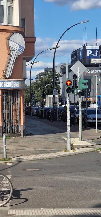Straßenszene in Berlin-Wedding mit Fußgängerampel auf Rot, einem großen Schlüsselschild an einem Eckhaus und parkenden Autos entlang der Straße.

