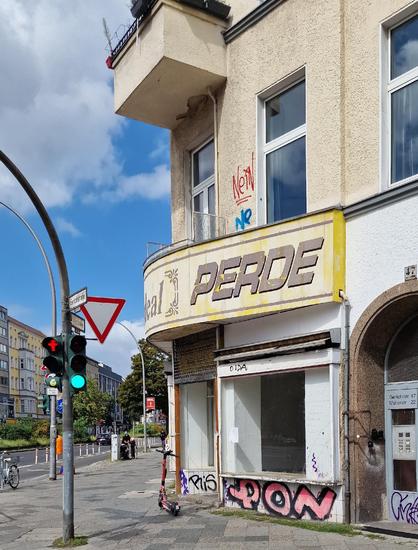 Alt-Text: Straßenecke in Berlin-Wedding mit einem leerstehenden Ladengeschäft, über dessen Eingang ein vergilbtes Schild mit der Aufschrift „Perde“ hängt; Fassade mit Graffiti, daneben eine Ampel und ein abgestellter E-Scooter.

