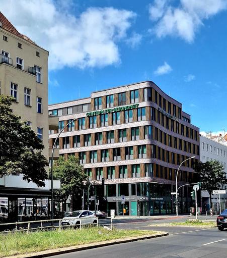 Das Bild zeigt ein modernes Eckgebäude in einer Stadt. Auf der Fassade ist in großen Buchstaben „Centrum für Gesundheit“ zu lesen, im Erdgeschoss befindet sich eine Filiale der Krankenkasse AOK. Das Gebäude hat eine geschwungene Form mit abwechselnden Elementen aus Glas und Holzverkleidung, die den Fenstern Struktur geben. Es hebt sich architektonisch deutlich von den älteren Nachbarhäusern ab. Vor dem Haus verlaufen Straßen mit Autos und Ampeln, daneben ist eine kleine Grünfläche mit Bäumen zu…