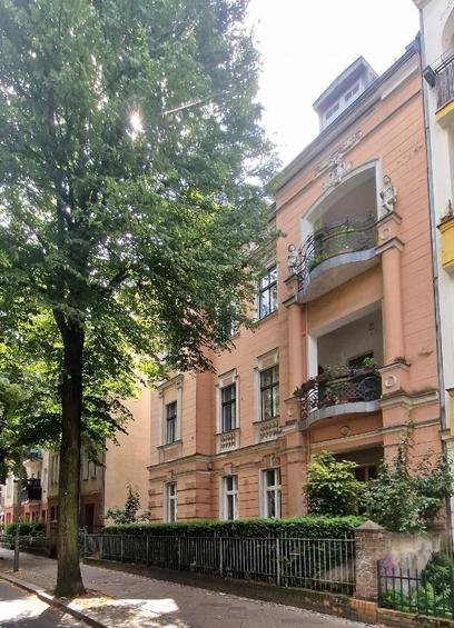 Das Foto zeigt ein Wohngebäude im Altbau-Stil mit einer reich verzierten Fassade in warmen Pfirsichtönen. Auffällig sind die Balkone mit geschwungenen schmiedeeisernen Geländern sowie Figuren und dekorative Elemente über den Eingängen. Das Haus wirkt historisch und gepflegt, es ist von einem Zaun und Grünpflanzen umgeben. Links im Bild verläuft eine ruhige Straße mit großen, dicht belaubten Bäumen, die Schatten spenden und dem Umfeld eine grüne, angenehme Atmosphäre verleihen.

