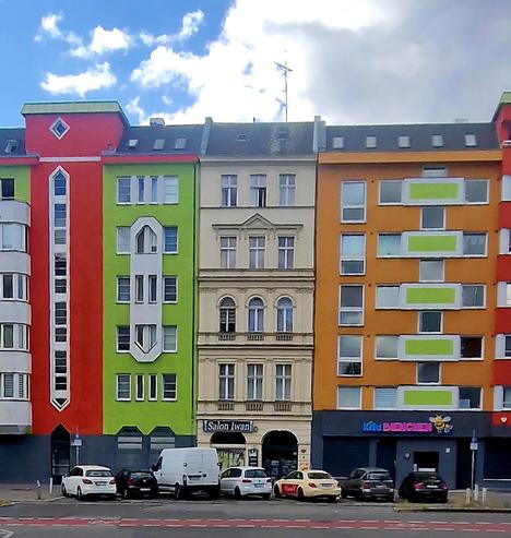 Straßenansicht mit nebeneinander stehenden Wohnhäusern in Wedding: Ein farbiges Ensemble mit orange, grün und rot gestrichenen Fassaden sowie einem älteren, beigefarbenen Altbau in der Mitte. Im Erdgeschoss befinden sich Geschäfte, davor sind mehrere Autos geparkt.

