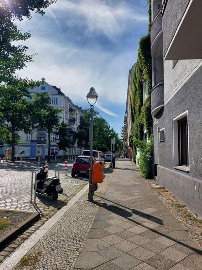 Straßenszene in Berlin-Wedding mit Kopfsteinpflaster, Gehweg, parkenden Autos und einem Motorroller; rechts Häuserfassaden, teilweise mit Efeu bewachsen, in der Mitte eine Straßenlaterne mit orangem Mülleimer.

