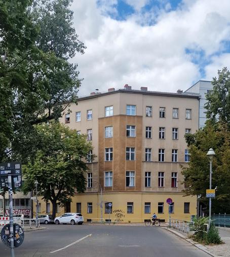 Mehrstöckiges Wohnhaus an einer Straßenecke in Berlin-Wedding, mit beigefarbener und ockerfarbener Fassade, Bäumen im Vordergrund und einem Radfahrer auf der Straße.

