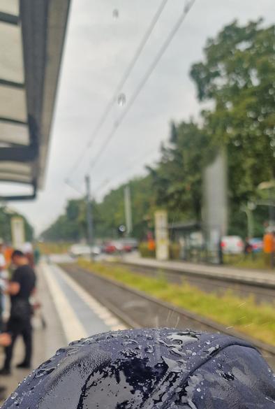 Alt-Text: Nasse blaue Kappe mit Regentropfen im Vordergrund, im Hintergrund ein Bahnsteig mit Schienen, wartenden Personen und Bäumen.

