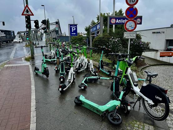 Mehrere umgestürzte und abgestellte E-Scooter sowie ein E-Bike von Lime liegen und stehen ungeordnet auf einem nassen Gehweg an einer Kreuzung in Berlin-Wedding, im Hintergrund eine Brücke und Verkehrsschilder.

