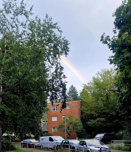 Gehweg mit parkenden Autos, Fahrrädern und einem E-Scooter, im Hintergrund ein Backsteinwohnhaus und ein Regenbogen am Himmel.

