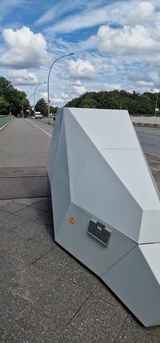 Graues, kantiges Metallgehäuse mit Blitzer auf einem Gehweg an einer Straße mit Laternen und Bäumen im Hintergrund.

