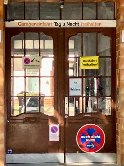 Braune Holztüren mit Glasfenstern, darauf mehrere Schilder mit Parkverbot und dem Hinweis „Garageneinfahrt Tag u. Nacht freihalten“.

