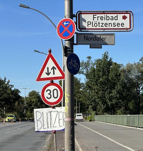 Straßenszene am Nordufer in Berlin-Wedding mit mehreren Verkehrsschildern, darunter Tempo-30-Zone, Vorfahrtsregelung und Radweg, sowie Wegweiser zum Freibad Plötzensee; handgemaltes Schild „Blitzer“ ist ebenfalls angebracht.

