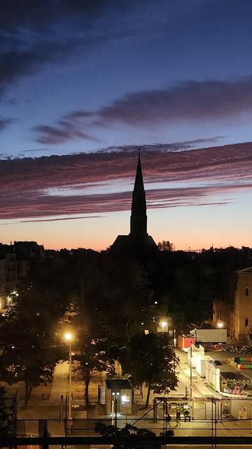 Alt-Text: Silhouette einer Kirche mit spitzem Turm bei Sonnenuntergang in Berlin-Wedding, im Vordergrund ein beleuchteter Platz mit Bäumen und Buden.

