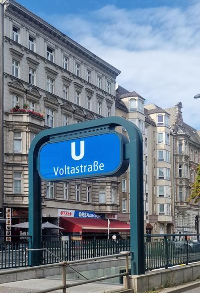 Blaues U-Bahn-Schild mit der Aufschrift „Voltastrße“ vor mehrstöckigen Altbauten in Berlin-Wedding.

