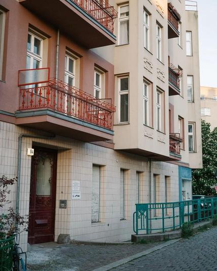 Dieses Foto zeigt ein Wohngebäude mit mehreren Etagen und Balkonen. Die Fassade ist in einem hellen Beige- und rötlich-braunen Ton gehalten. Die Balkone sind mit verzierten roten Geländern ausgestattet. Im Erdgeschoss befinden sich geschlossene Rollläden vor den Fenstern sowie eine dunkelrote Eingangstür mit dekorativen Schnitzereien.
Neben der Tür hängt die Hausnummer 64, außerdem sind dort einige Schilder angebracht. Entlang des Gehwegs verläuft ein grünes Metallgeländer, und der Boden ist m…