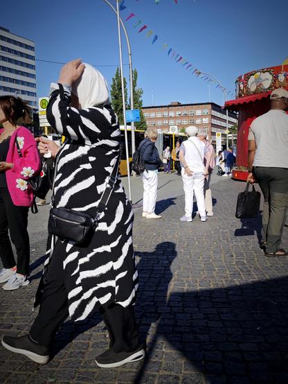 Alt-Text: Menschen auf dem Leopoldplatz in Berlin-Wedding bei sonnigem Wetter, mit Bussen im Hintergrund, bunten Wimpeln über dem Platz und einem roten Karussellrand rechts im Bild.

