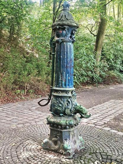 Alt-Text: Historische gusseiserne Wasserpumpe mit verzierten Reliefs und blauer Lackierung, auf einem gepflasterten Platz im Grünen.

