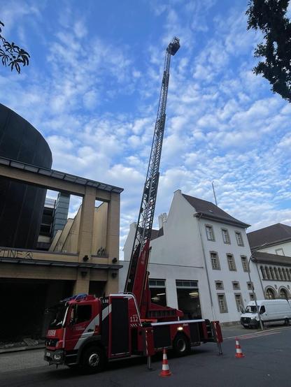 Alt-Text: Feuerwehrfahrzeug mit ausgefahrener Drehleiter, abgesperrt mit Leitkegeln, vor Gebäuden in einer Straße.

