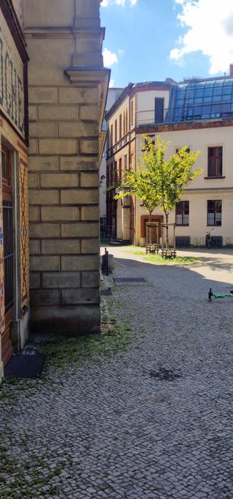 Alt-Text: Gepflasterter Innenhof in Berlin-Wedding mit alten Backsteingebäuden, einem jungen Baum in der Mitte und einem abgestellten E-Scooter am rechten Bildrand.

