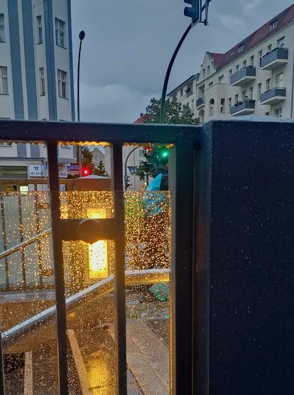 Alt-Text: Nasse Glasfläche mit Regentropfen am Eingang einer U-Bahn-Station in Berlin-Wedding, dahinter beleuchtete Treppe, Straßenlaternen und Wohnhäuser bei Abenddämmerung.

