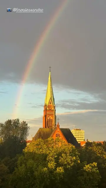 Kirchturm der Neuen Nazarethkirche  in Berlin-Wedding mit Regenbogen am Himmel, umgeben von Bäumen im Vordergrund.

