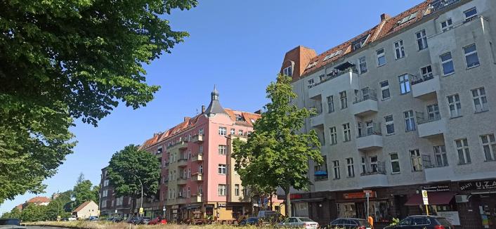 Straßenszene in Berlin-Wedding mit mehrstöckigen Wohnhäusern, einem rosafarbenen Eckhaus mit Turmspitze und Geschäften im Erdgeschoss, davor Bäume und parkende Autos.

