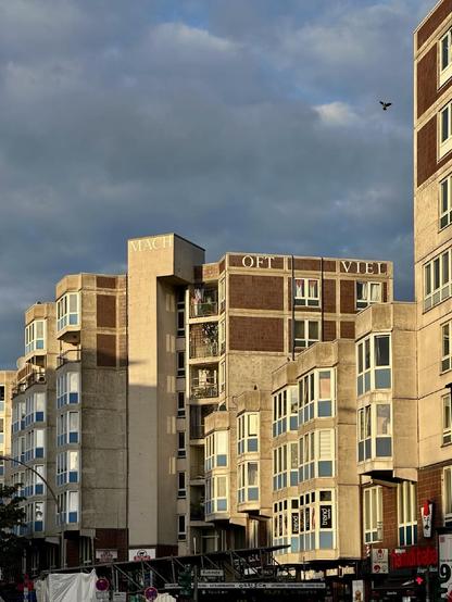 Wohnblock im Berliner Wedding mit markanten Vorbauten und den großen weißen Schriftzügen „MACH OFT VIEL“ auf dem Dachrand, im Vordergrund Straßenschilder und Geschäfte.

