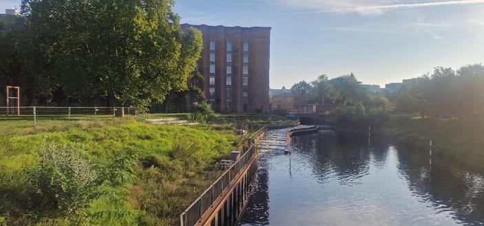 Backsteingebäude an einem Kanal in Berlin-Wedding, links eine Grünfläche mit Bäumen und Bauzäunen, rechts Wasser mit kleiner Staustufe.

