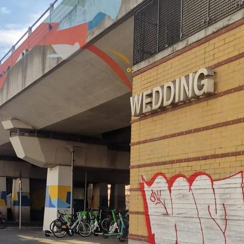 Alt-Text: Gelbes Backsteingebäude mit der Aufschrift „WEDDING“ am Bahnhof Wedding, darunter Fahrräder und E-Scooter; im Hintergrund eine Brücke mit bunter Wandbemalung.

