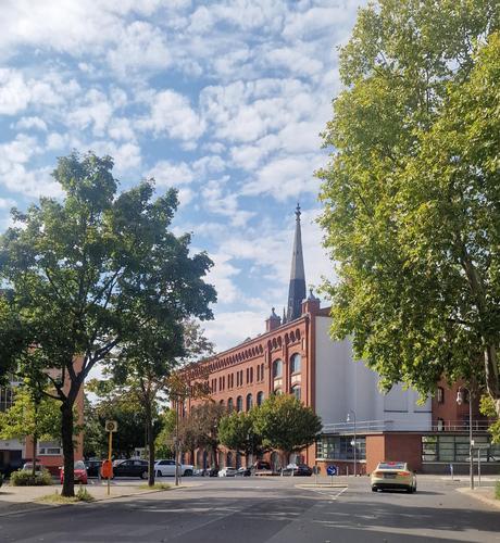 Alt-Text: Rote Backsteinfassade eines historischen Gebäudes mit hohem Kirchturm in Berlin-Wedding, umgeben von Bäumen und parkenden Autos an einer Straßenkreuzung.

