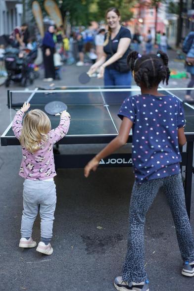 Kinder spielen auf einer für den Verkehr gesperrten Straße im Berliner Stadtteil Wedding bei einer Aktion der temporären Spielstraße; zu sehen sind unter anderem Tischtennis, Laufräder, Straßenmalkreide, Roller und ein großes buntes Schwungtuch.