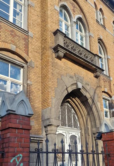 Backsteinfassade eines historischen Gebäudes in Berlin-Wedding mit Spitzbogenportal, verzierten Fenstern und steinernem Balkon; Baujahr „1887–1888“ ist über dem Eingang eingraviert.