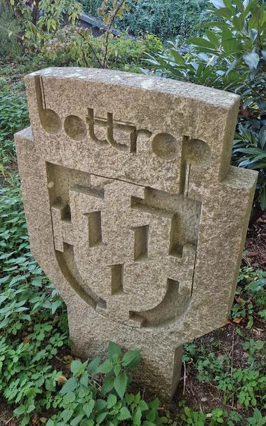 Steinerne Stele mit der eingravierten Aufschrift „Bottrop“ und einem stilisierten Wappen, umgeben von Grünpflanzen.

