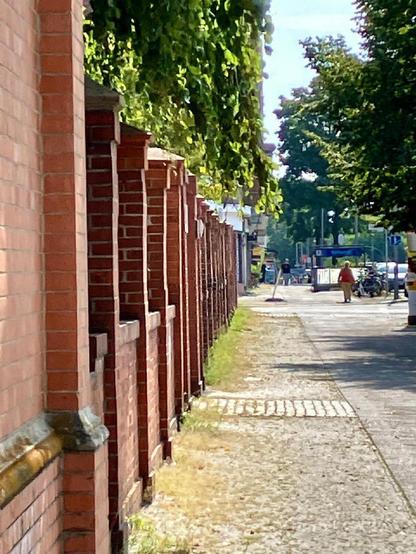 Rote Backsteinmauer mit vorspringenden Pfeilern entlang eines Gehwegs in Berlin-Wedding, im Hintergrund Bäume und eine Straße mit Passanten.

