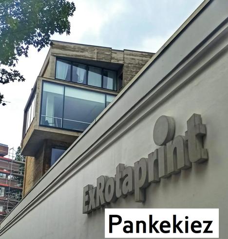 Auf dem Foto ist das Eingangsschild von ExRotaprint zu sehen, einem bekannten Kultur- und Gewerbestandort im Berliner Wedding. Das Gebäude mit seinen großen Glasfronten im Hintergrund ist Teil des sanierten Geländes der ehemaligen Druckmaschinenfabrik. Unten im Bild ist zusätzlich der Schriftzug „Pankekiez“ eingefügt, der auf das umliegende Quartier entlang der Panke verweist.