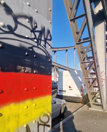Alt-Text: Metallkonstruktion einer Brücke in Berlin-Wedding mit Graffiti in Schwarz und den Farben der Deutschlandflagge, daneben parkende Autos und ein weißer Lieferwagen.

