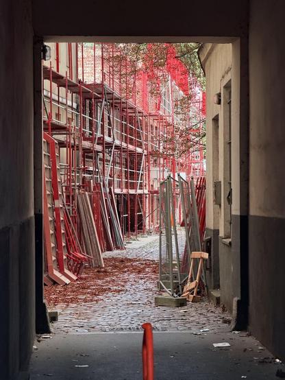 Alt-Text: Blick durch einen Torbogen auf einen Innenhof mit Kopfsteinpflaster, in dem rote Baugerüste und Metallteile gelagert sind.

