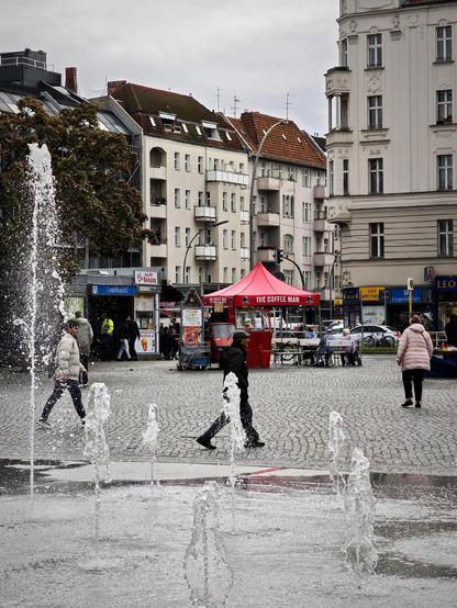 Springbrunnen auf dem Leopoldplatz in Berlin-Wedding, mit Passantinnen und Passanten auf dem gepflasterten Platz, einem roten Kaffeestand und umliegenden Wohnhäusern im Hintergrund unter grauem Himmel.

