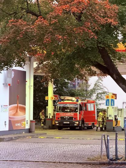 Ein rotes Feuerwehrfahrzeug steht an einer Shell-Tankstelle in Berlin-Wedding. Mehrere Feuerwehrleute in Schutzkleidung arbeiten an den Zapfsäulen, während herbstlich gefärbte Bäume den Ort überschatten.

