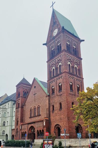 Backstein-Kirchgebäude mit hohem Turm und grünem Dach in Berlin-Wedding, neugotischer Stil mit Rundbogenfenstern und Uhr unter dem spitzen Dach. Vor dem Gebäude stehen Straßenschilder und ein Baum mit Herbstlaub.

