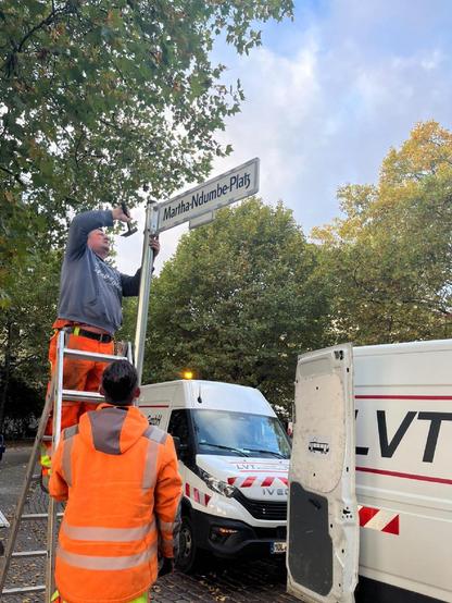 Zwei Arbeiter in orangefarbener Arbeitskleidung montieren ein Straßenschild mit der Aufschrift „Martha-Ndumbe-Platz“ an einem Pfosten. Einer steht auf einer Leiter, während der andere unten hilft. Im Hintergrund stehen ein weißer Transporter und herbstliche Bäume.

