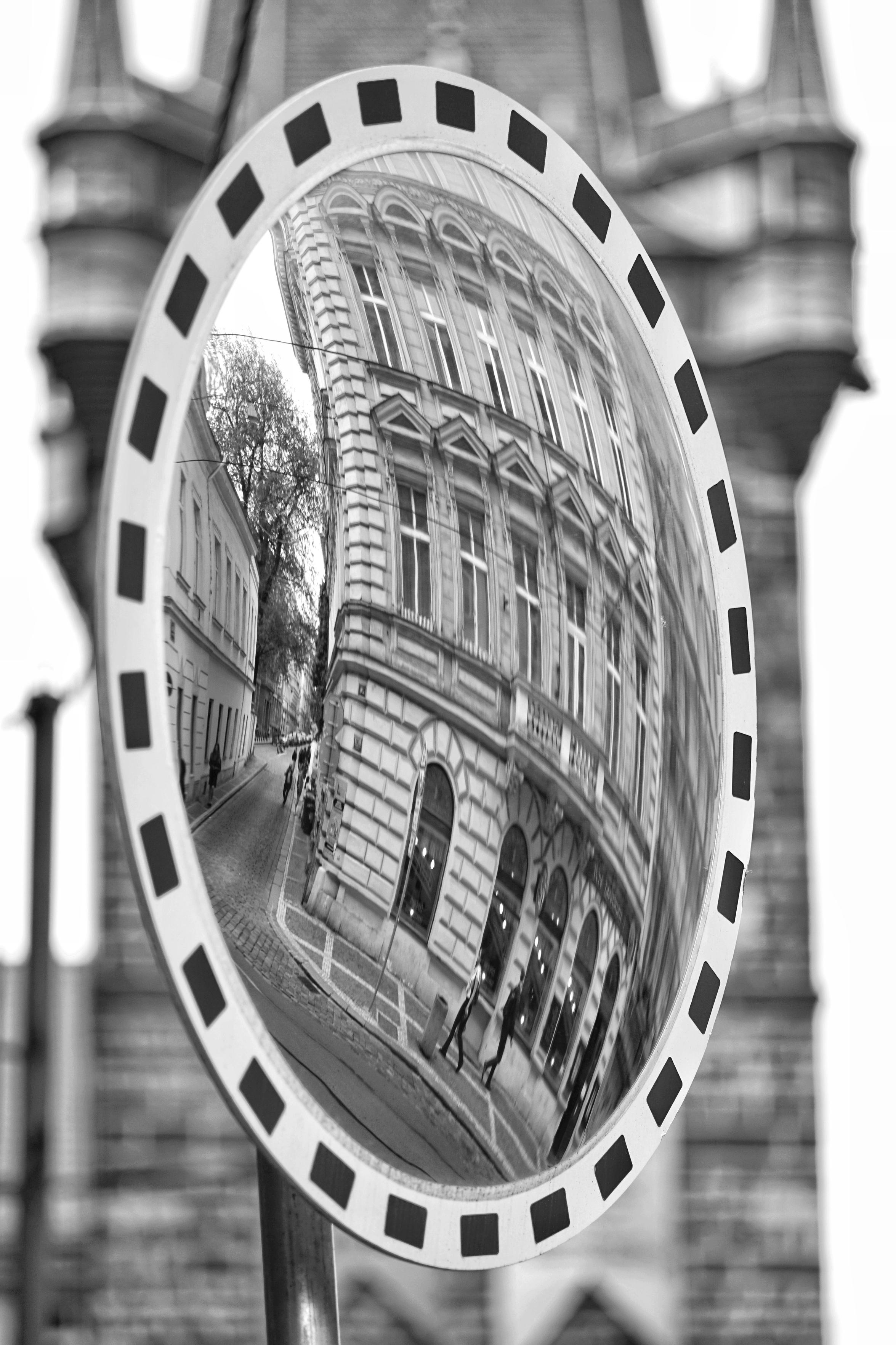 Schwarz-Weiß-Foto im Hochformat. Zu sehen ist ein Verkehrsspiegel, in dem sich ein Jugendstilgebäude spiegelt. Hinter dem Spiegel steht ein Turm, er ist unscharf zu erkennen.