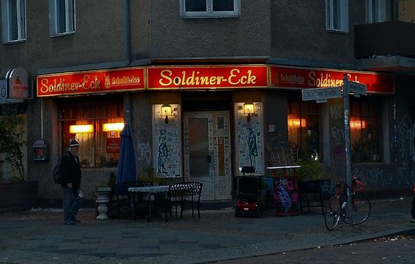 Eckkneipe mit rotem Leuchtschild „Soldiner-Eck“ an der Ecke einer Straße im Berliner Stadtteil Wedding, mit beleuchteten Fenstern, Graffiti an der Fassade, einem Fahrrad und einem Mann auf dem Gehweg.