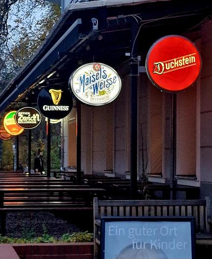 
„Außenbereich eines Lokals mit überdachten Sitzbänken; mehrere beleuchtete runde Biermarken-Schilder hängen an der Fassade, darunter Duckstein, Maisel’s Weisse, Guinness und Zwiefalter; im Hintergrund geht eine Person entlang, vorne steht ein Werbeschild.“

