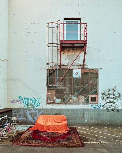Abgenutzte Hausfassade mit großem Industrie­fenster und rotem Metallpodest; davor ein mit orangefarbenem Tuch bedecktes Sofa auf einem Teppich, daneben ein Fahrrad und Graffiti an der Wand.