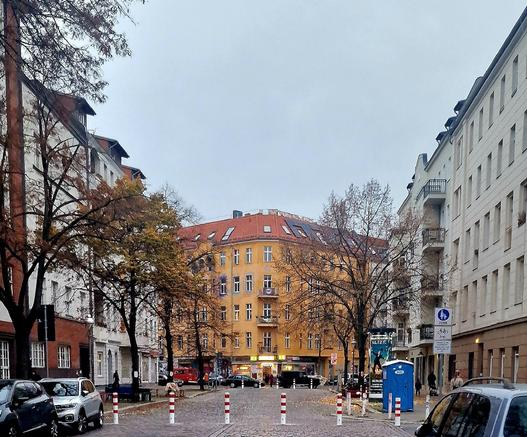 Kopfsteinpflasterstraße im Wedding mit mehrgeschossigen Wohnhäusern, gelbem Eckhaus im Zentrum, herbstlichen Bäumen und parkenden Autos; weiße Poller sperren die Durchfahrt, rechts steht eine blaue mobile Toilette.