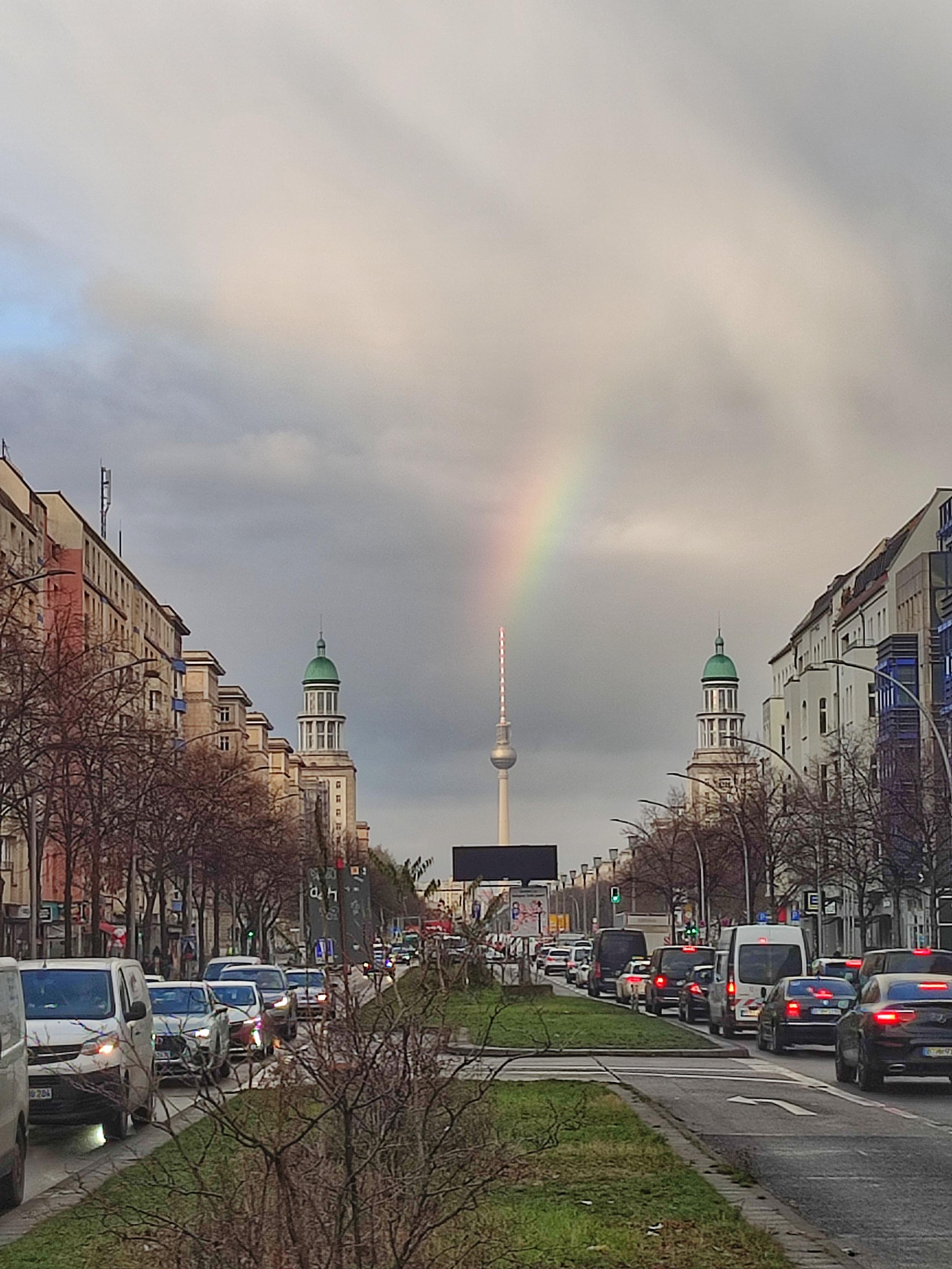 Blick auf die Frankfurter Allee Richtung Berliner Fernsehturm. Rechts und links Häuser und Autos, im Hintergrund die Türme am Frankfurter Tor und der Fernsehturm. Am Himmel ein Regenbogen, er sieht aus, als käme er direkt aus der Spitze des Fernsehturms.