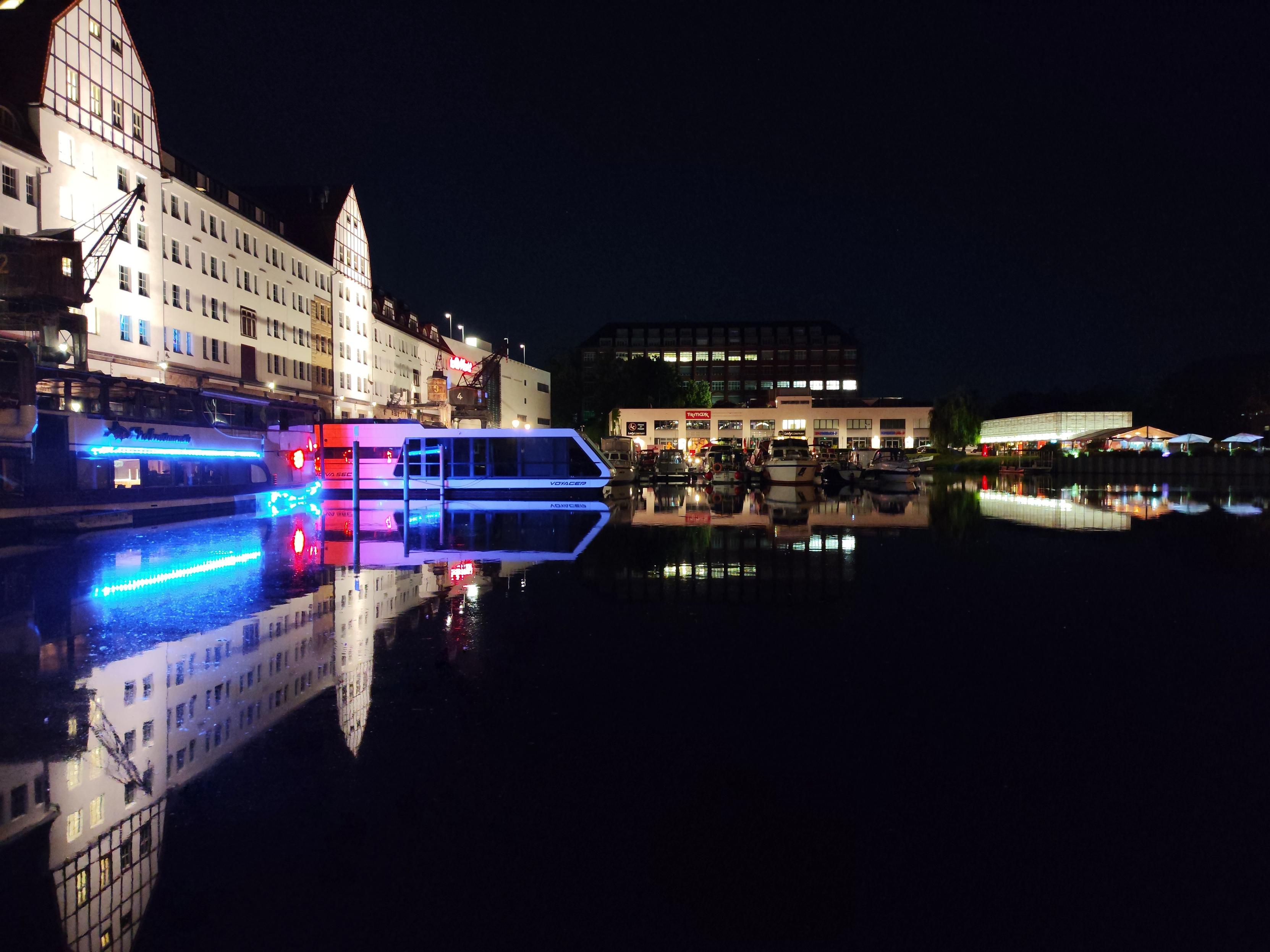 Farbfoto im Querformat. Zu sehen sind beleuchtete Hause und Schiffe an einem Hafen, sie Siegeln sich im Wasser. Von links nach rechts sind immer weniger und niedrigere Motive im Bild, so dass das gesamte Motiv fast wirkt wie ein Pfeil nach rechts.